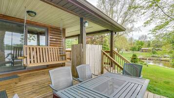 A covered outdoor deck with wooden furniture, a hanging swing, and a view of a pond and trees.