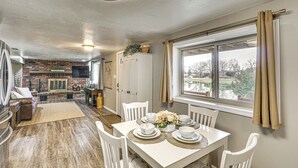 Un comedor con una mesa blanca dispuesta para cuatro personas, rodeada de sillas blancas. En el fondo, una chimenea de ladrillo, y una ventana con cortinas a la derecha.
