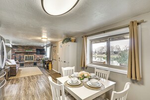 Un comedor con una mesa blanca dispuesta para cuatro personas, rodeada de sillas blancas. En el fondo, una chimenea de ladrillo, y una ventana con cortinas a la derecha.