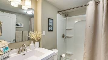 Une salle de bain avec un lavabo blanc, un miroir et une douche. On y trouve des serviettes et des articles de toilette sur le lavabo.