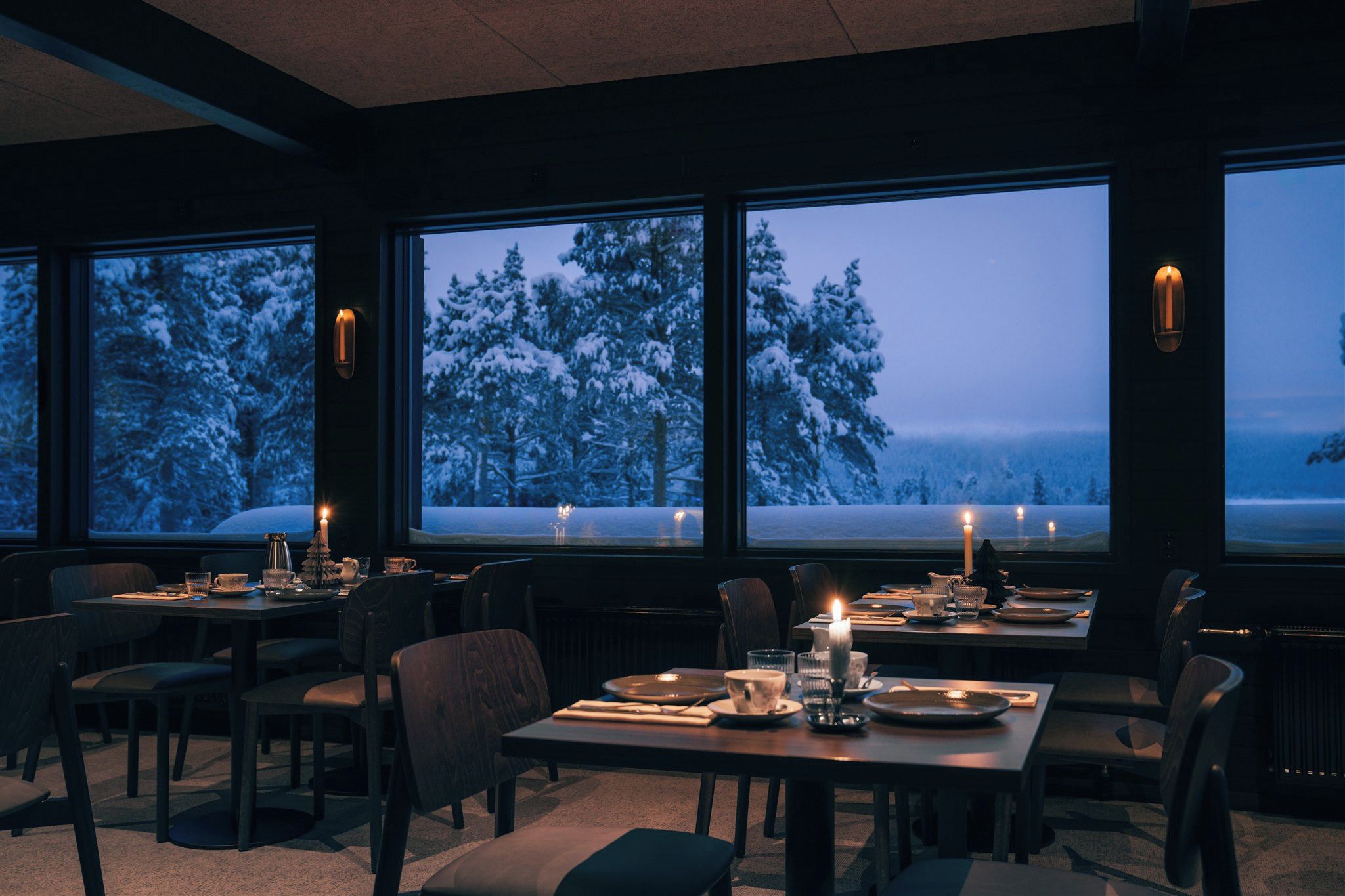 A dining area with tables set for two, large windows with a snowy landscape view, and candles on the tables.