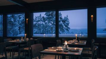 Uma sala de jantar com mesas preparadas para dois, grandes janelas com vista para uma paisagem coberta de neve e velas sobre as mesas.