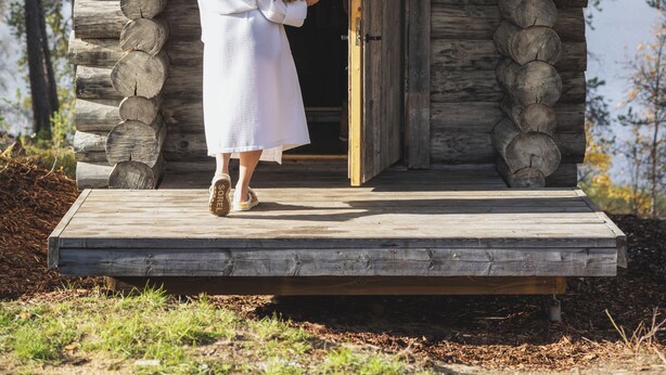 Une cabane en bois avec un porche, une personne debout à l’entrée et un lac en arrière-plan.