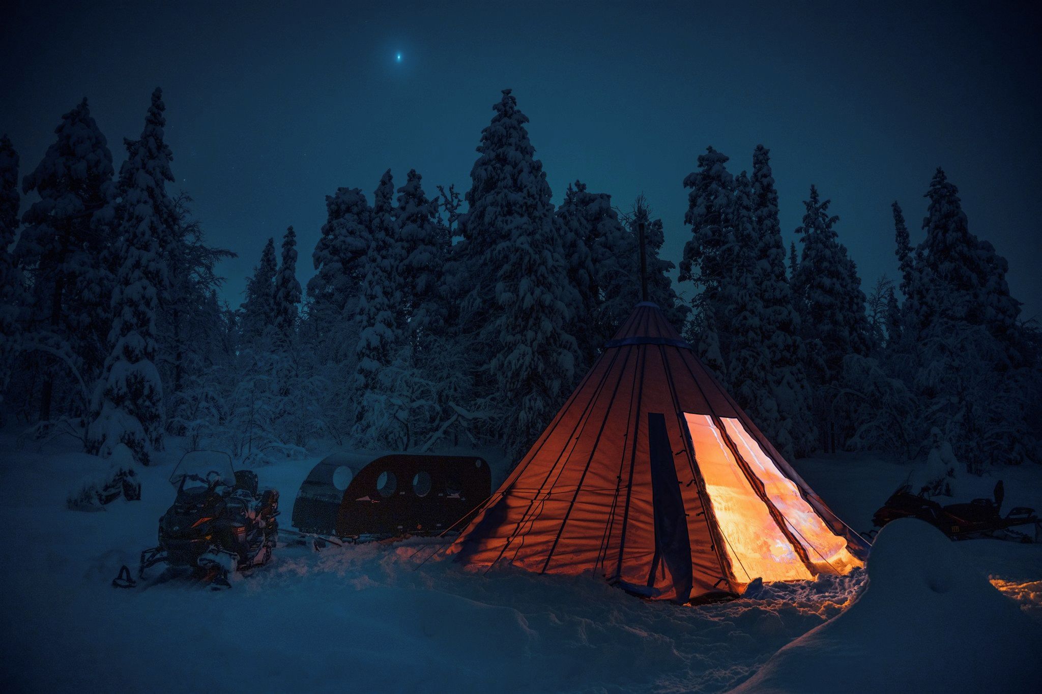 A snow-covered forest with a tent illuminated by a fire, a snowmobile, and a vehicle in the background.