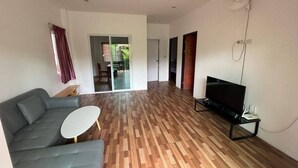 A modern living room with a grey sofa, a small white table, and a TV on a black stand. The room has wooden flooring and a ceiling fan. - Spacious 2bdr BangTao House  (Choeng Thale)