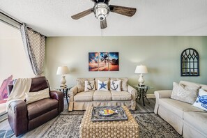A living room with a brown leather sofa, beige armchairs, a coffee table with a decorative fish, and a ceiling fan.