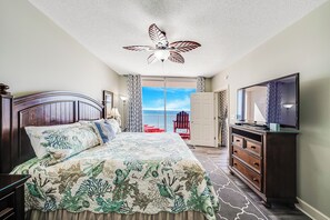 Une chambre avec un lit, un ventilateur de plafond, une télévision, une commode, une porte donnant sur un balcon et une vue sur le ciel.
