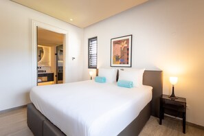 Habitación de hotel moderna con una cama grande, una mesita de noche con lámpara, un cuadro enmarcado en la pared y un baño visible a través de una puerta abierta.