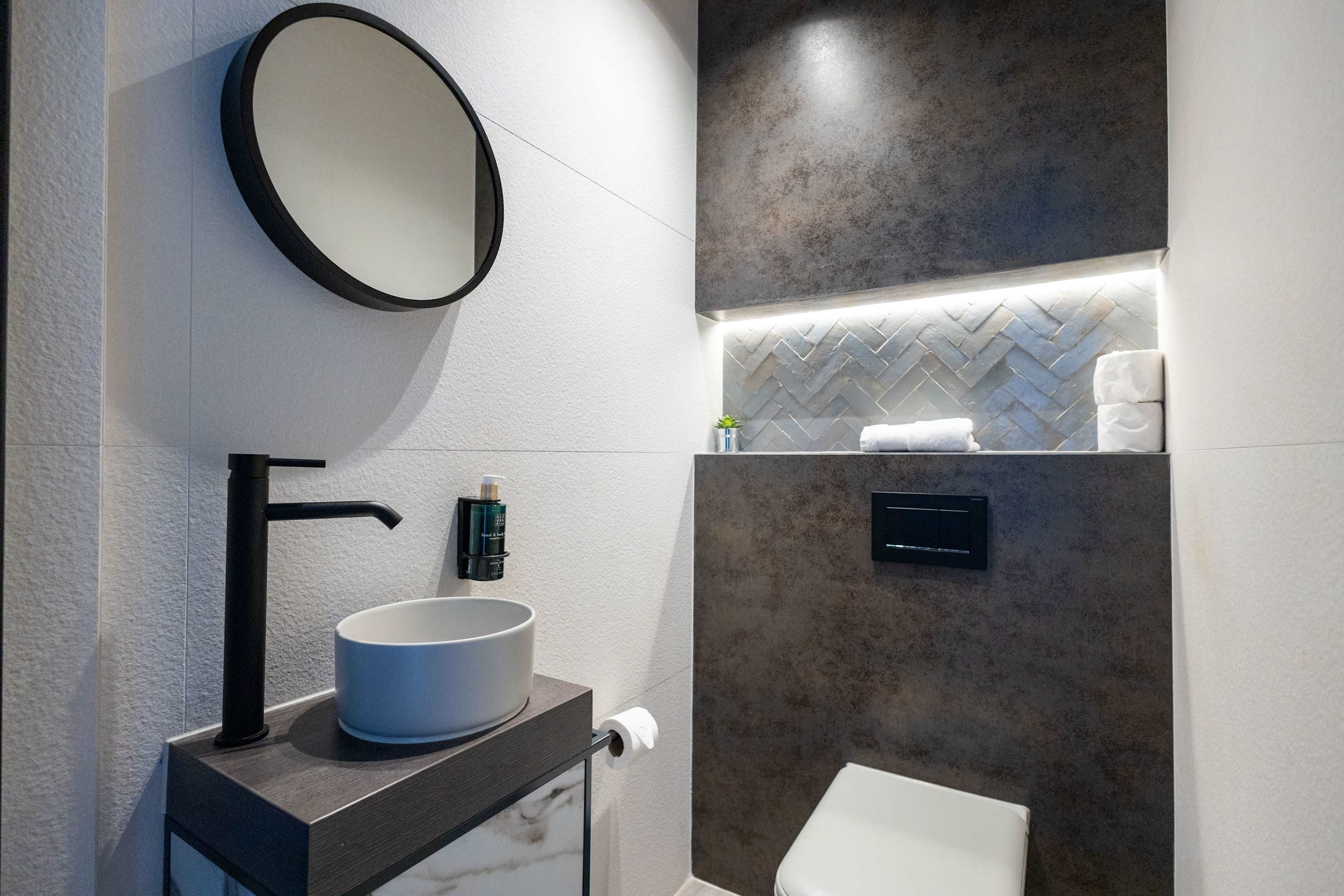 A modern bathroom with a round mirror, a black faucet, a white sink, and a toilet.