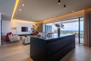Une cuisine moderne dotée d’un grand îlot central, d’un téléviseur à écran plat et offrant une vue sur la mer grâce à de grandes fenêtres.