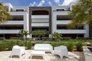 Un hôtel moderne à l’architecture contemporaine, doté d’une structure à plusieurs étages, d’un espace extérieur aménagé avec des meubles en plastique blanc et d’un jardin paysager avec différentes espèces de plantes.
