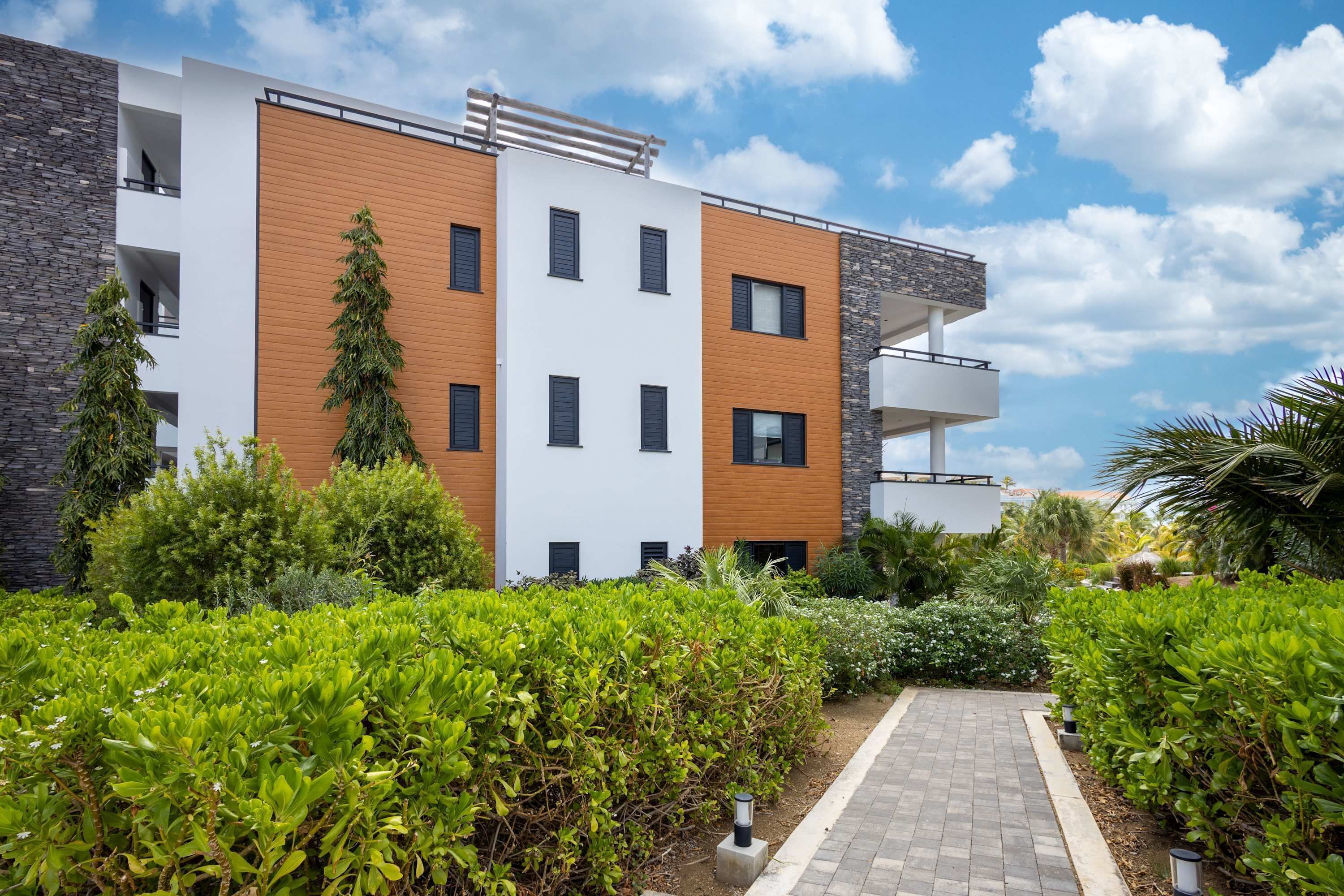 A modern multi-story building with a mix of white and wooden facades, surrounded by well-maintained greenery and a paved pathway.