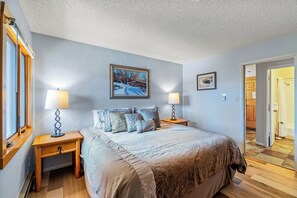 A bedroom with a large bed, two bedside tables with lamps, a framed picture on the wall, and a wooden door leading to another room.
