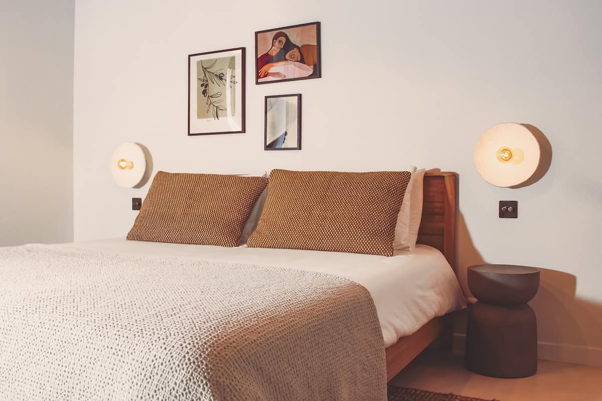 Une chambre à coucher bien rangée, avec un lit, des oreillers, des lampes fixées au mur et des œuvres d’art encadrées.