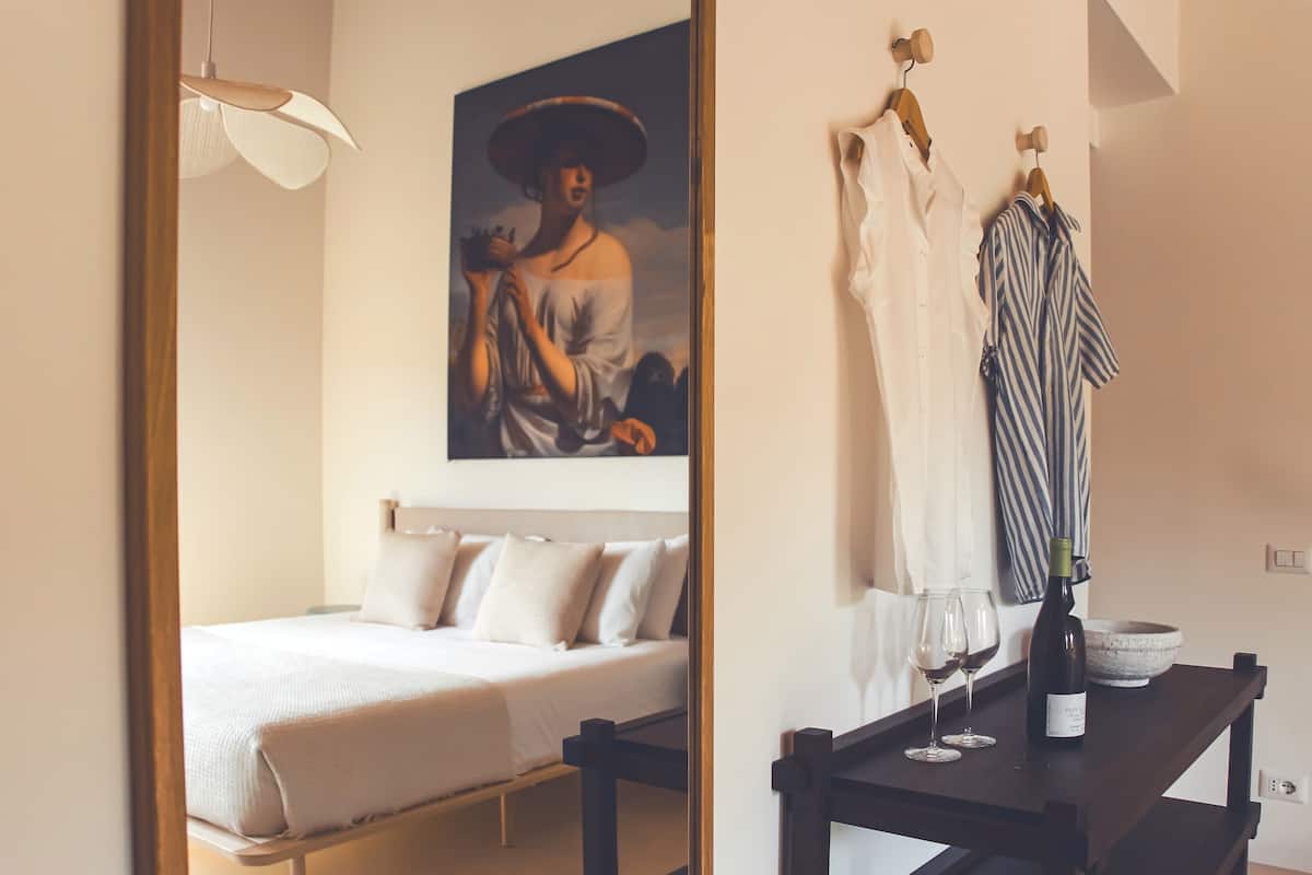 Une chambre à coucher avec un lit, un tableau, une table avec des verres à vin et une bouteille, et des vêtements suspendus.