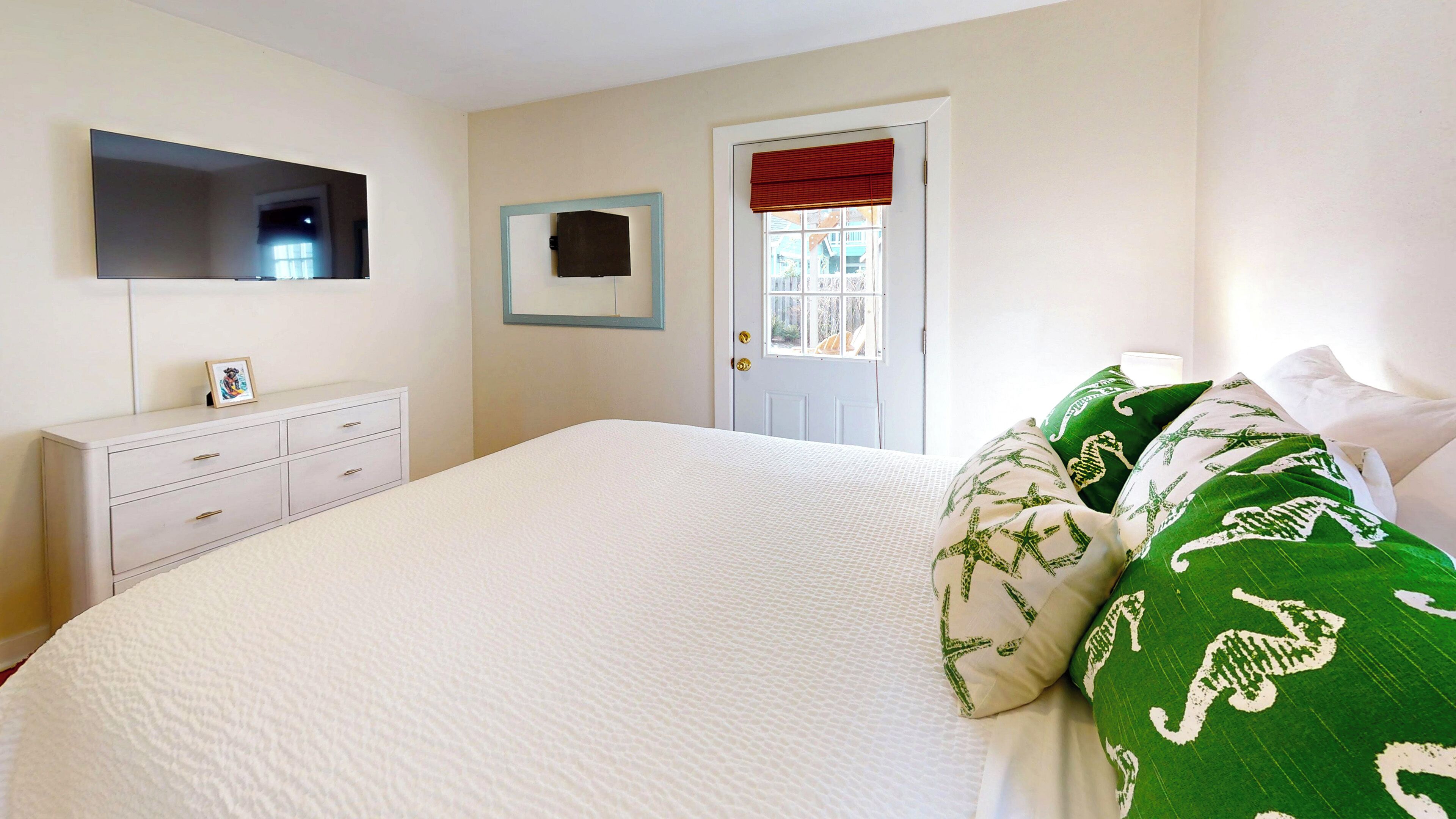 Une chambre avec un grand lit, une commode à tiroirs, un téléviseur à écran plat fixé au mur et une porte avec une fenêtre.