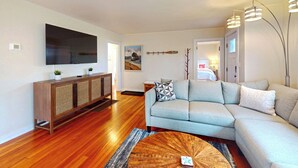 A living room with a sectional sofa, a wooden cabinet with woven doors, a flat-screen TV, and a painting of Oregon. - Georgie's Tide and True Cottage at Seaside Beach (Seaside)