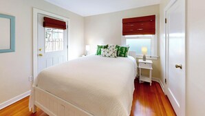 A bedroom with a bed, bedside tables, a nightstand, a window with blinds, and a door. - Georgie's Tide and True Cottage at Seaside Beach (Seaside)