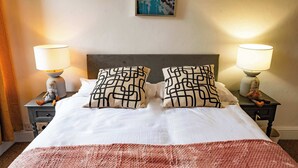 Ein ordentlich bezogenes Bett mit gemusterten Kissen, zwei Nachttischlampen und einem gerahmten Kunstwerk an der Wand.