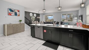 A modern kitchen with a central island, stainless steel appliances, and black cabinetry. There are two pendant lights, a staircase with a railing, and a view of the outdoors through a window.