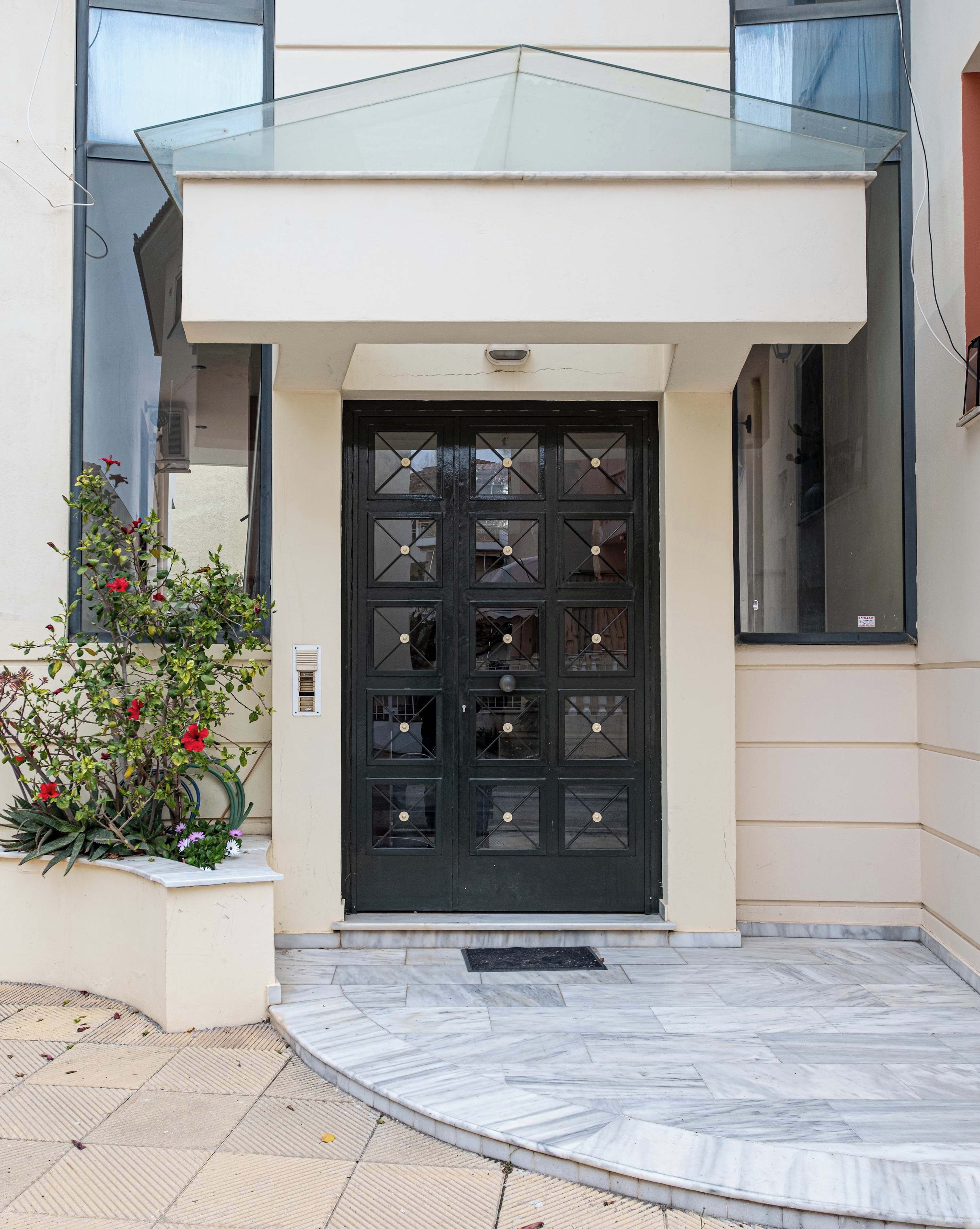 A modern building entrance with a black door, a glass awning, and a small garden with red flowers.