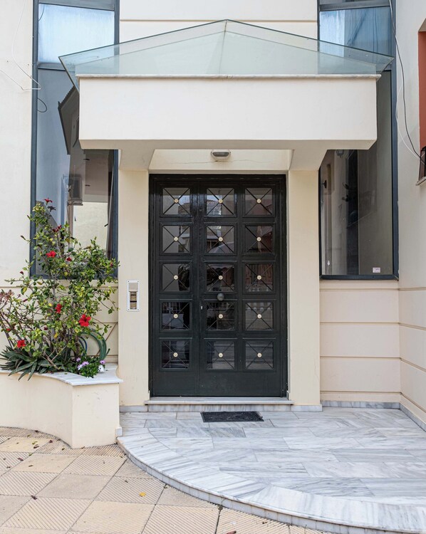 A modern building entrance with a black door, a glass awning, and a small garden with red flowers.