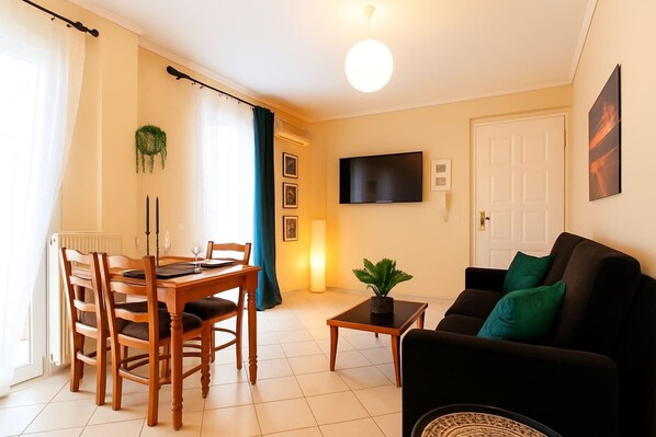 A living room with a black sofa, a dining table with chairs, a TV mounted on the wall, and a door with a window.