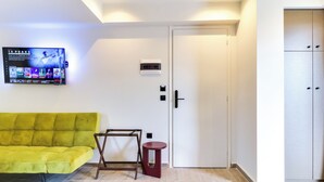 Une pièce avec un canapé vert, un petit tabouret rouge, un téléviseur fixé au mur, une porte avec une poignée et une armoire avec un miroir.