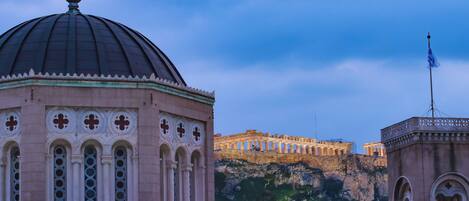 A historical building with a dome and arched windows, a flagpole, and a view of ancient ruins on a hill.