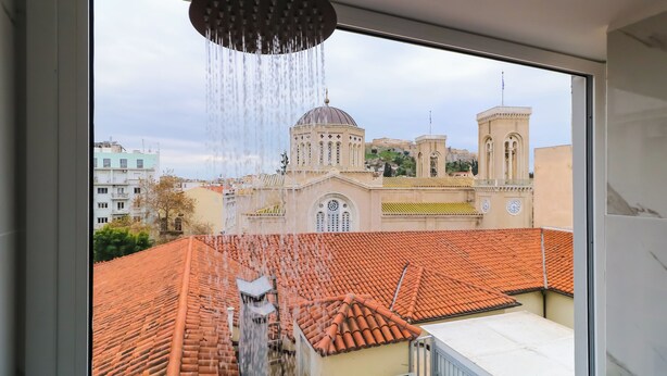 Une vue depuis une fenêtre, avec un pommeau de douche en évidence, donnant sur un bâtiment doté d’un dôme et d’une tour.