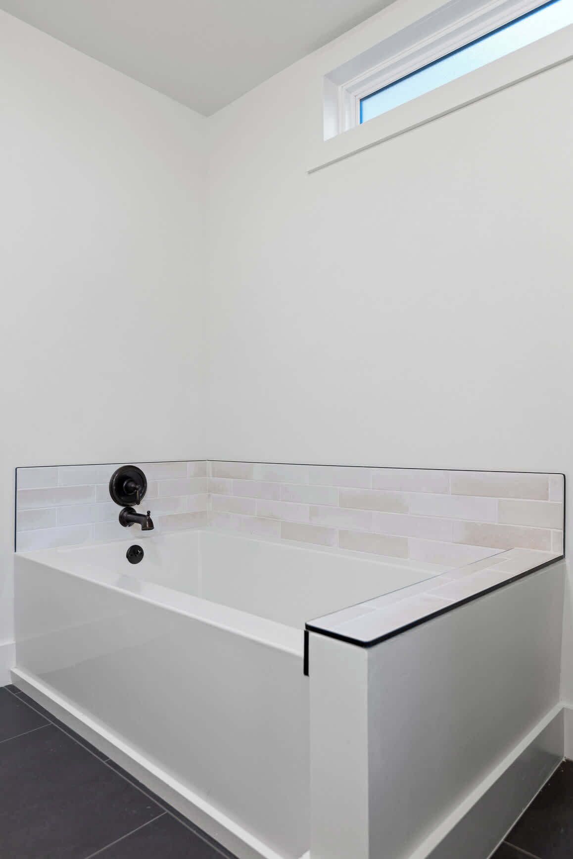 A modern bathroom with a white bathtub, a small window above it, and a tiled floor.
