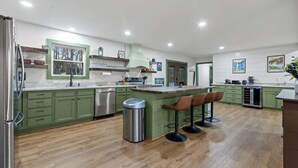 Una cuina moderna amb armaris verds, un refrigerador d'acer inoxidable, un rentaplat i un cubell de la brossa. Hi ha tamborets al taulell i una finestra sobre el rentaplat.