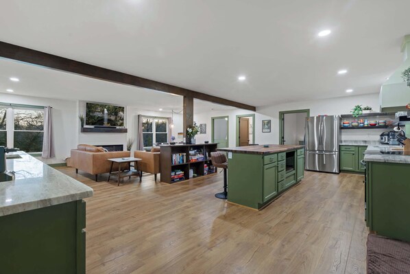 A spacious living area with a sofa and a coffee table, a kitchen with green cabinets and stainless steel appliances, and a large window providing natural light. - Hemlock Haven – Perfect for Groups of 16 (Pikeville)