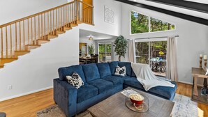 Un salon moderne avec un canapé d’angle bleu, un escalier en bois et de grandes fenêtres donnant sur l’extérieur.