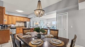 A dining area with a wooden table set for six, a chandelier, and a kitchen in the background. - San Remo 304 2BR Beachfront Condo (Redington Shores)