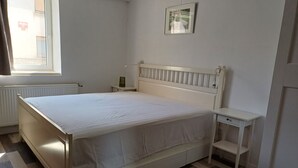 A bedroom with a bed, a bedside table, a nightstand, a radiator, a window, and a framed picture on the wall.