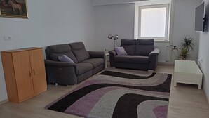 A modern living room with two grey sofas, a wooden cabinet, a floor lamp, a TV, and a window with blinds.