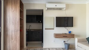 Un espacio habitable compacto con kitchenette, un televisor montado y un pequeño estante de madera.
