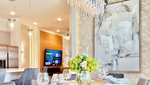 A modern dining area with a wooden table set for six, surrounded by grey upholstered chairs. A large abstract painting adorns the wall, and a flat-screen TV is mounted on the wall in the background. - Stylish Home,Themed Rooms, Pvt Pool, BBQ, Resort Amenities – Internacional Drive (Orlando)