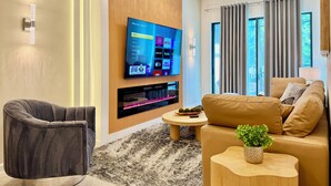 A modern living room with a flat-screen TV mounted on the wall, a comfortable sofa, a grey armchair, a small round coffee table, and a decorative plant. - Stylish Home,Themed Rooms, Pvt Pool, BBQ, Resort Amenities – Internacional Drive (Orlando)
