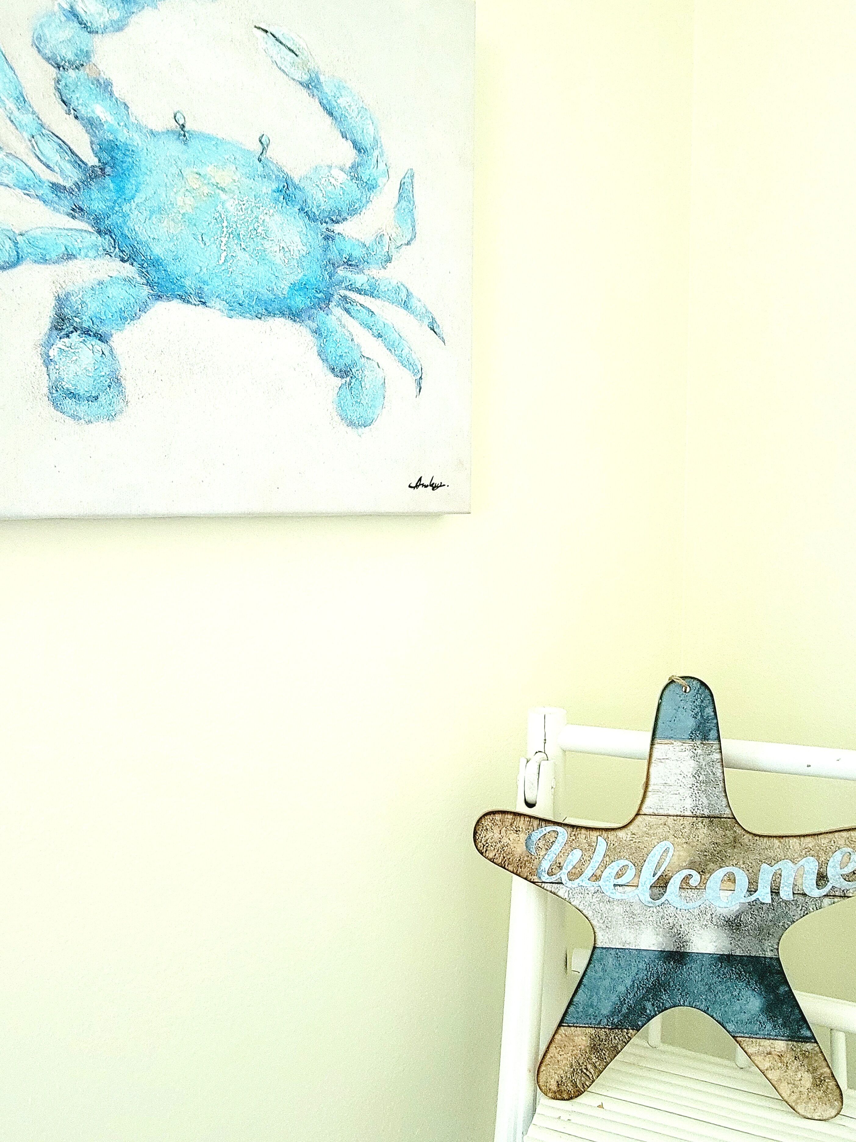 A wall with a blue crab painting and a welcome sign on a white chair.