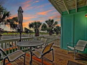 An outdoor patio with a round table and chairs, a wooden deck, and a view of a body of water and palm trees under a sunset sky.