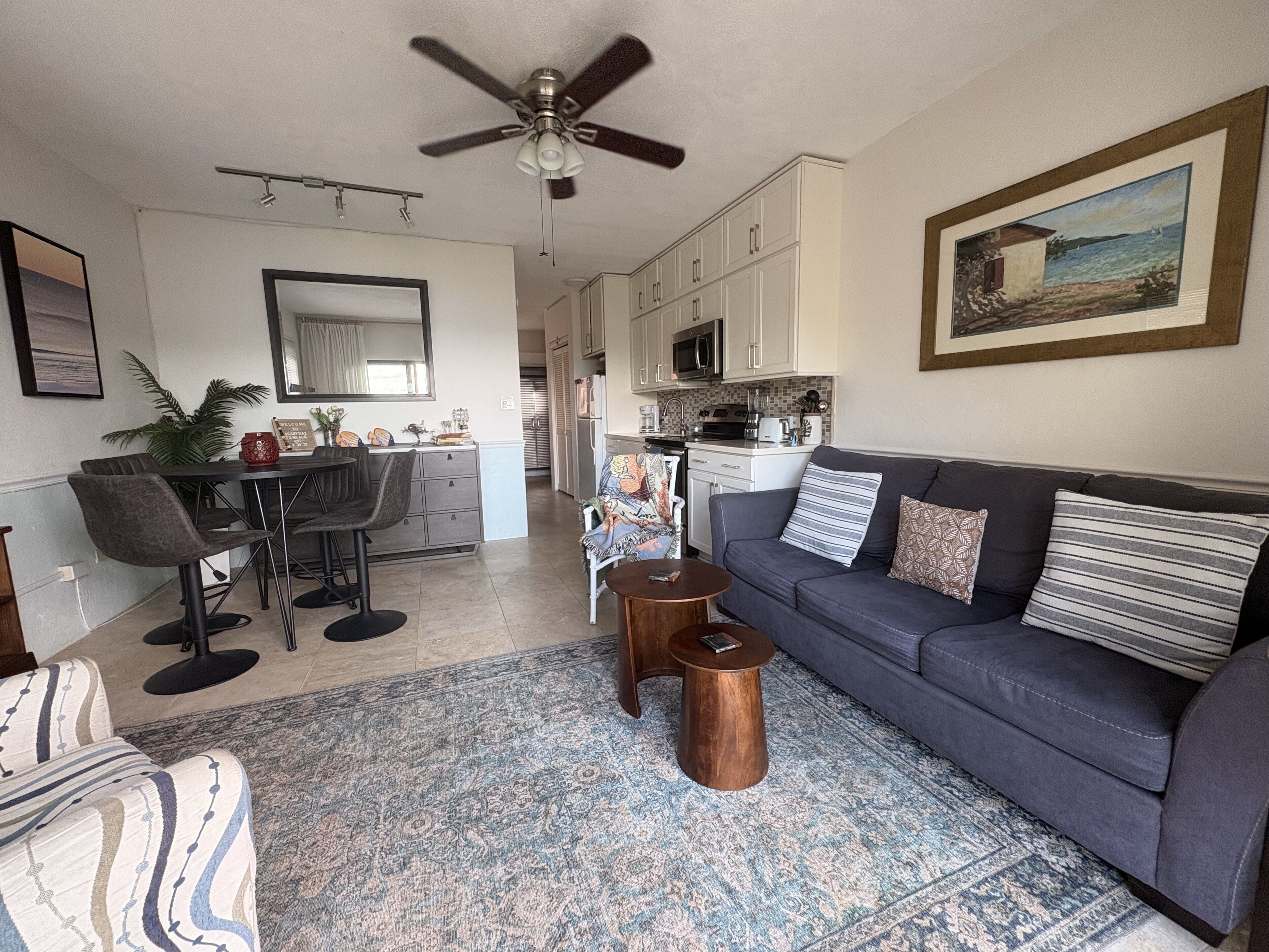 A living room with a sofa, coffee table, and chairs. There is a kitchen in the background with cabinets and appliances.