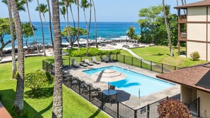 A pool area with lounge chairs and umbrellas, surrounded by palm trees and a view of the ocean. - Big Island Kona Isle E32 1 Bedroom Condo (Kailua-Kona)