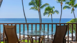 A balcony with two chairs and a glass table overlooking the ocean, with palm trees and a clear sky in the background. - Big Island Kona Isle E32 1 Bedroom Condo (Kailua-Kona)