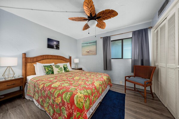 A bedroom with a large bed, a wooden headboard, a ceiling fan, a nightstand with a lamp, a chair, a window with curtains, and a picture of a beach on the wall.