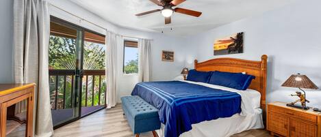 Un dormitorio con cama, un banco, una mesita de noche, un ventilador de techo y vistas a zonas verdes a través de una puerta corrediza.