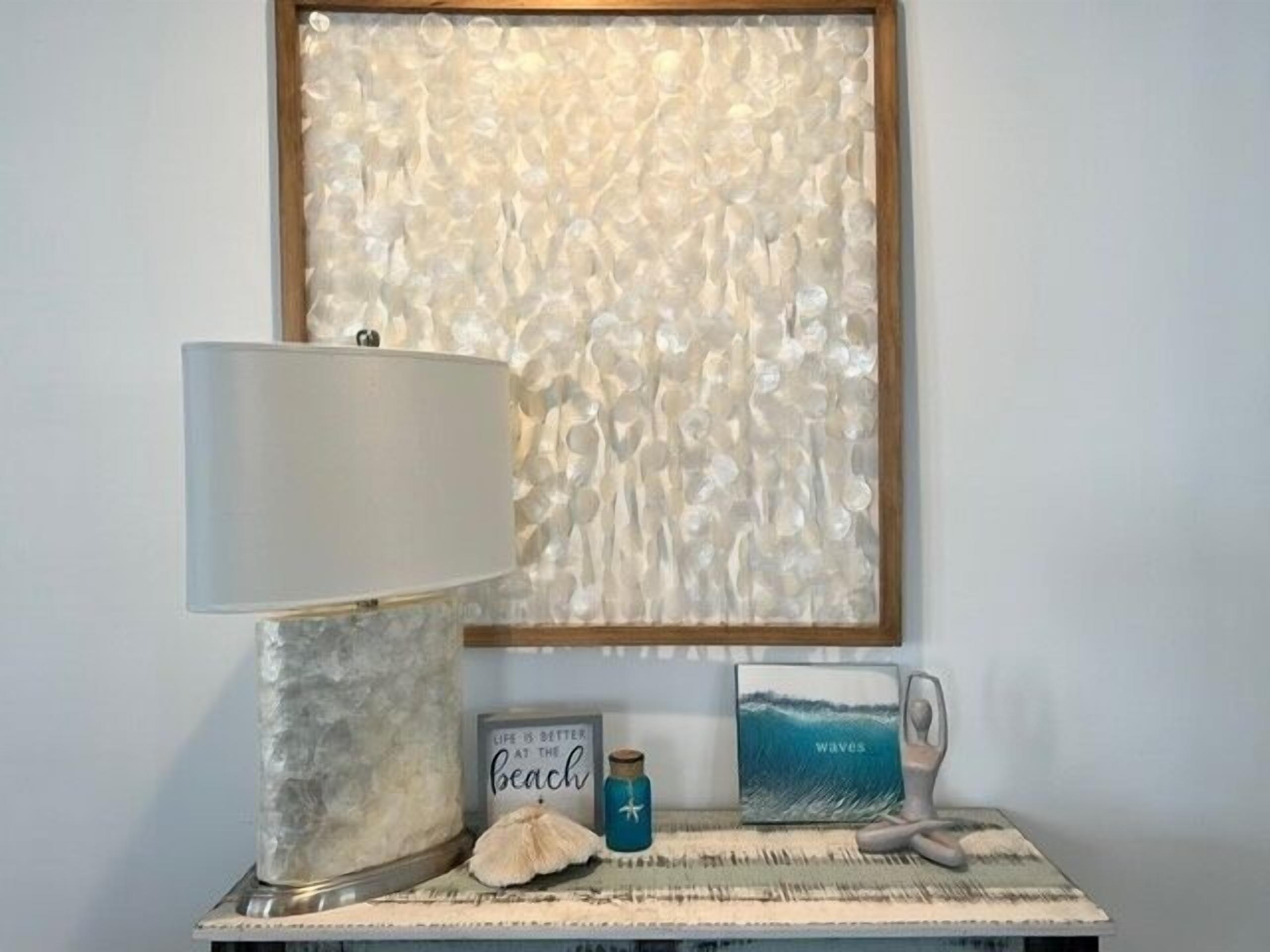 A room with a textured wall art, a lamp, a small table with a decorative item, a jar, and a framed picture.