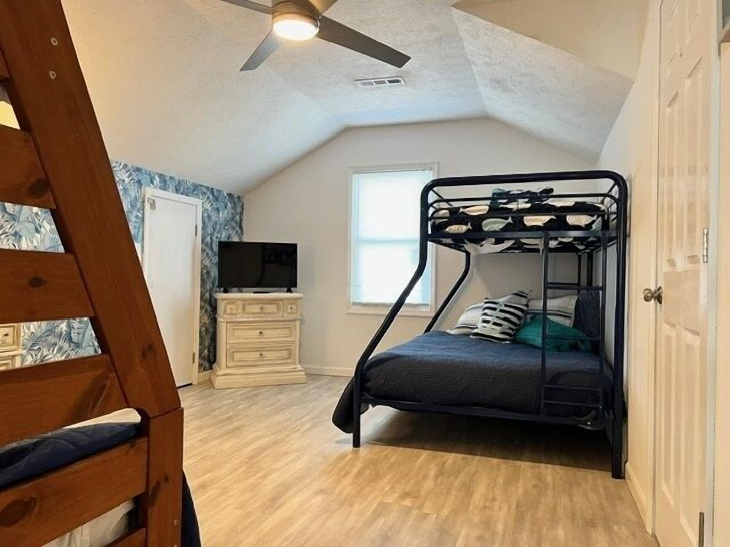 A bunk bed room with a ceiling fan, a dresser, a television, and a window.