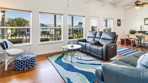 A spacious living room with a large gray sectional sofa, a white wicker chair, and a patterned ottoman. There are multiple windows offering views of the outside, and a ceiling fan is visible. - Bon Bini 4 Bedroom Home (Rockport)